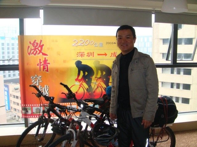 2008年李铁军站在两辆装有行李的自行车旁，背后展板写有2200公里深圳到成都。